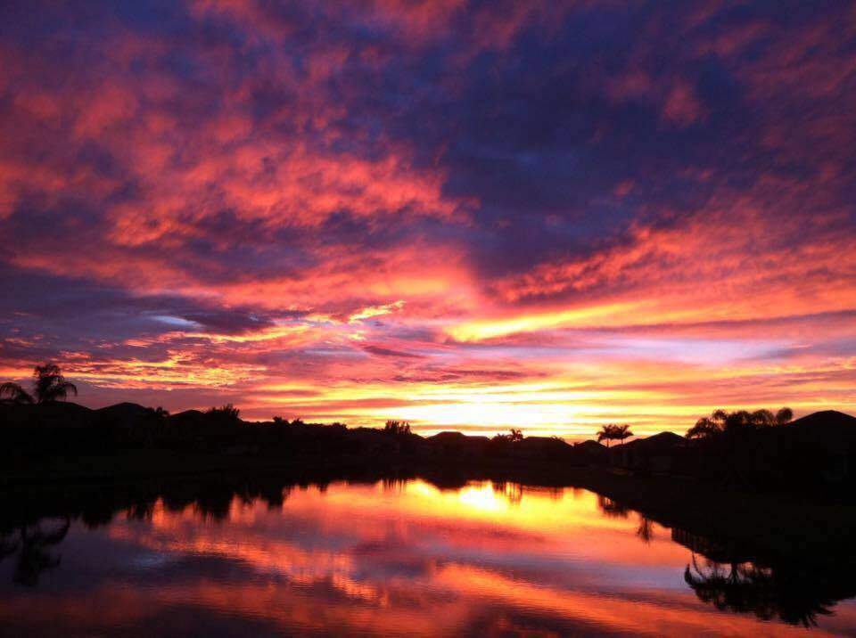 A vibrant sunset featuring a sky filled with shades of pink, orange, and purple is reflected in a calm body of water with silhouetted trees and houses along the horizon.