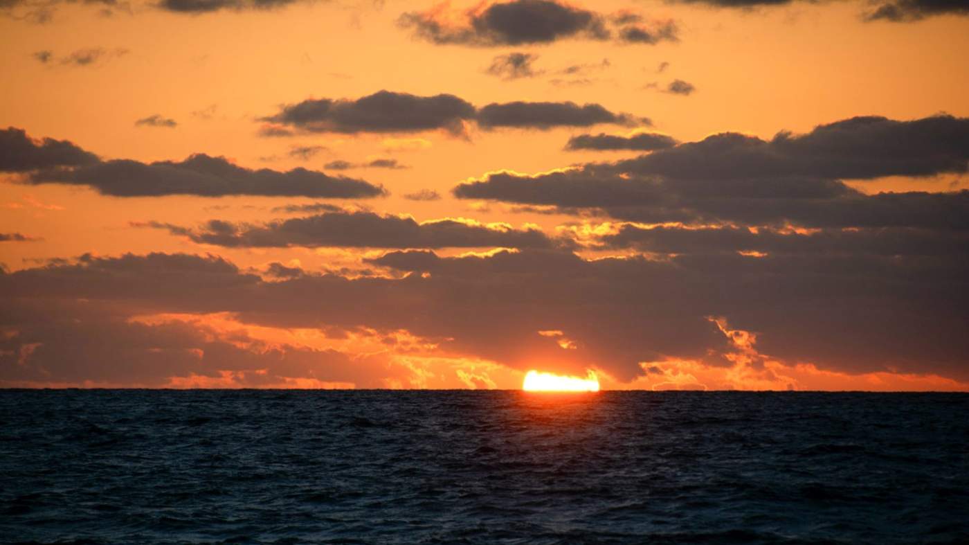 A serene sunrise over the ocean, with the sun partially dipped below the horizon. The sky displays shades of orange, yellow, and a hint of blue, while silhouetted clouds add contrast. The calm ocean reflects the sunrise’s colors.