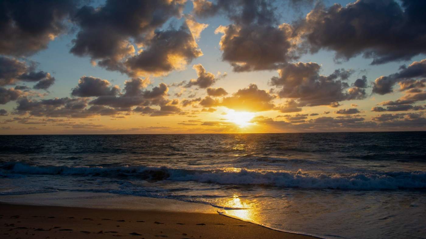 A tranquil beach at sunrise: The sun hangs low on the horizon, casting a warm glow across the sky and reflecting off the ocean. Fluffy clouds scatter across the sky, some partially blocking the sun’s rays, creating a dramatic effect. Gentle waves approach the sandy shore, where visible footprints lead toward the water. This picturesque scene captures nature’s beauty during the daily phenomenon of sunrise.