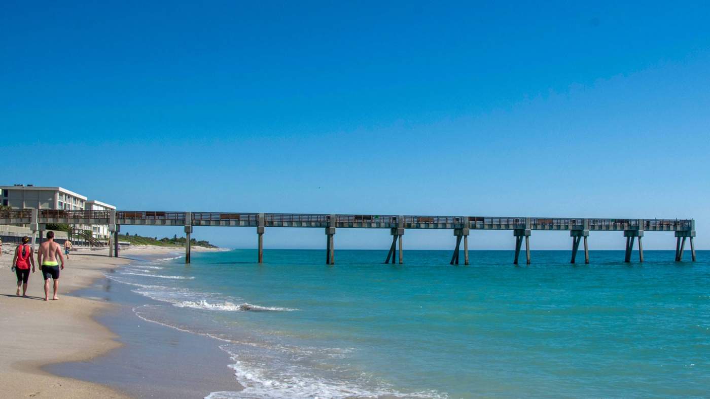 A clear day at the beach with a couple walking along the shoreline. The ocean is calm with gentle waves lapping at the sand. A long pier extends into the water on the right, and a building complex is visible on the left. The sky is a bright blue, indicating good weather