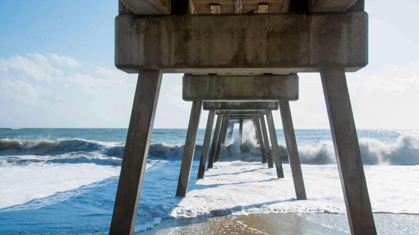 The image depicts the underside view of a pier extending the ocean. The concrete structure features vertical support columns and horizontal beams, creating a symmetrical pattern. Waves crash against the shore and pillars, with sunlight visible in the background.