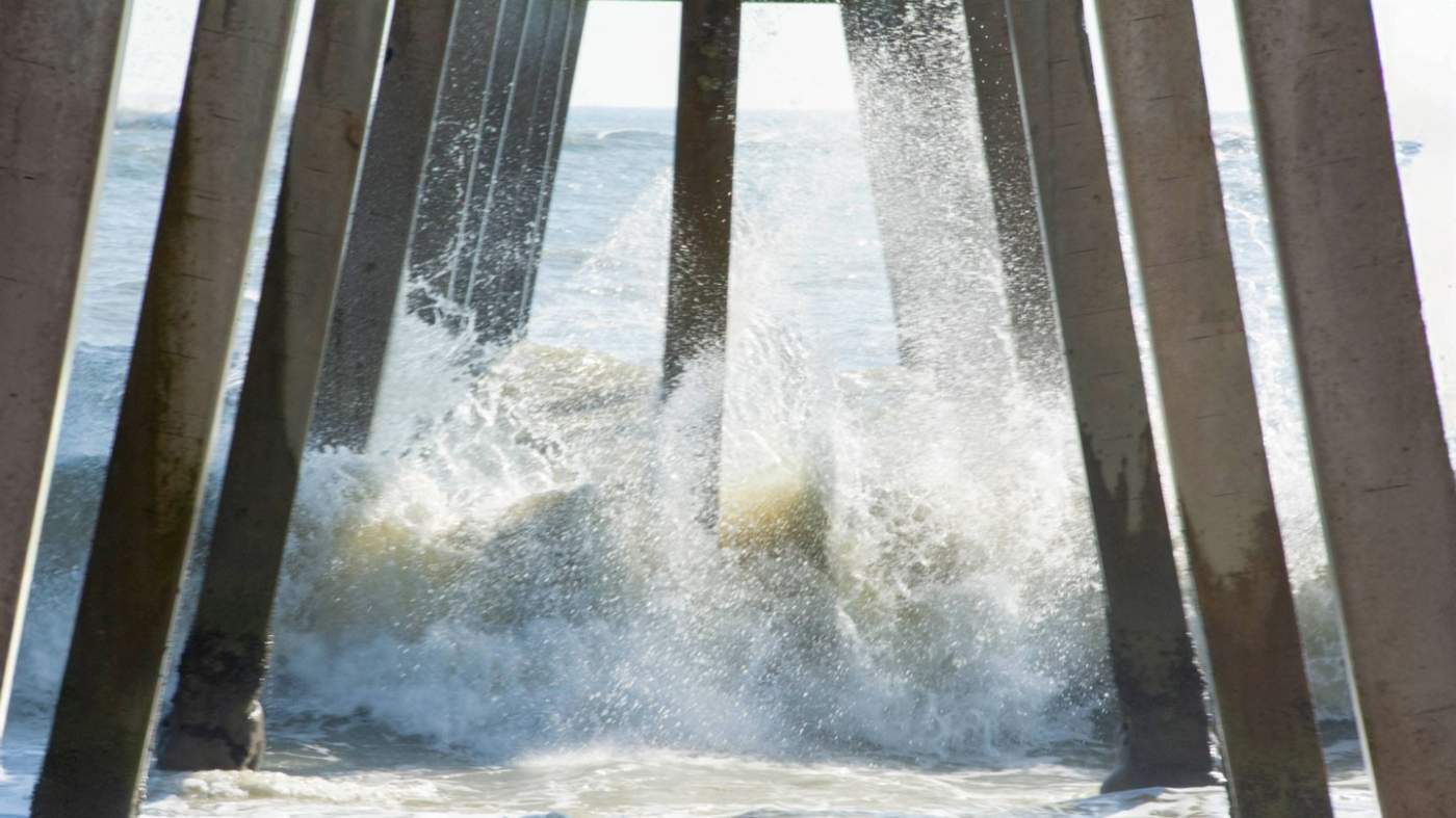 Sunlight filters through wooden pillars of a pier, waves crashing against them. The dynamic interaction between nature and man-made structure is captured, emphasizing the relentless force of water.”