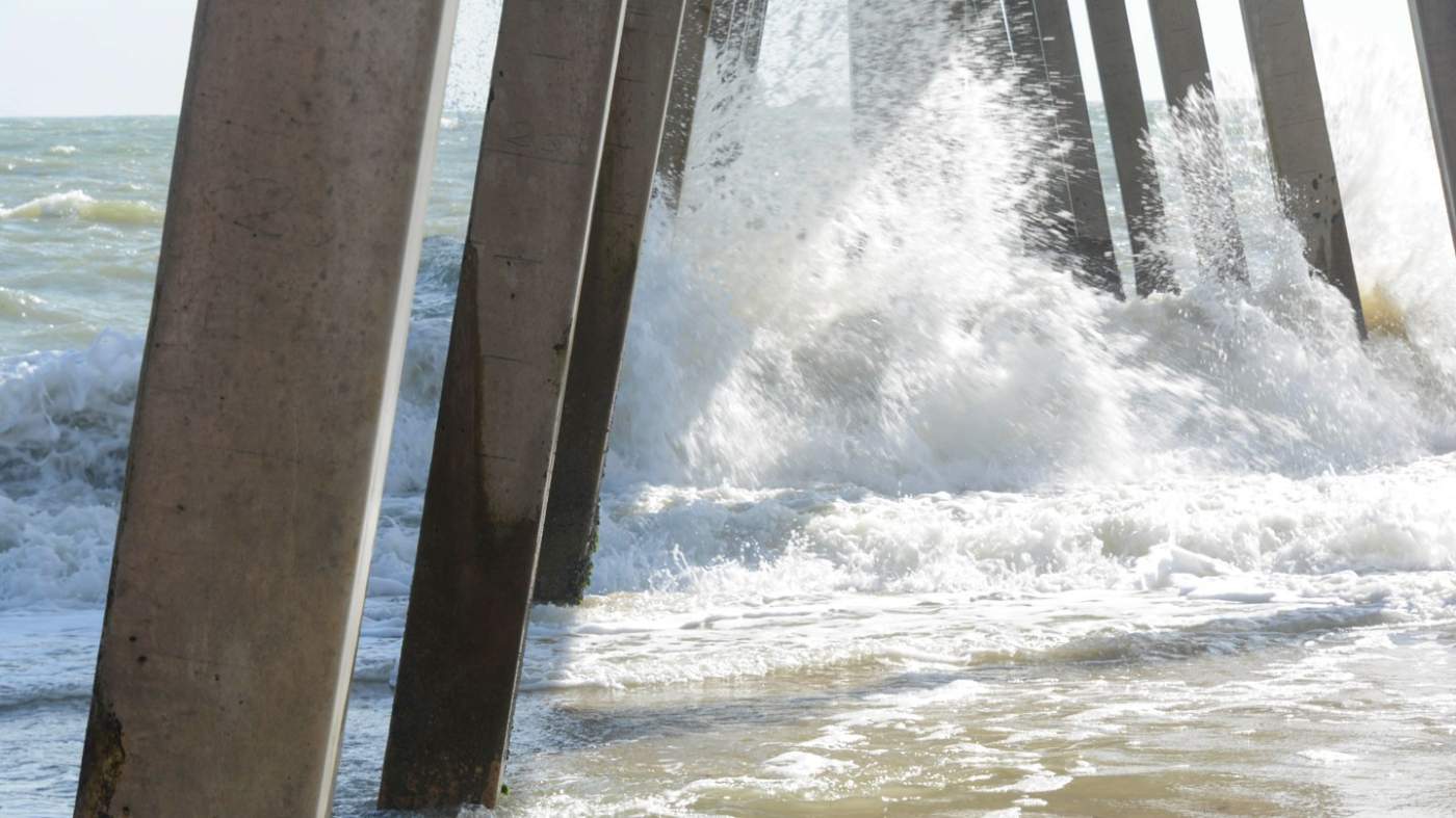 A dynamic scene of waves crashing against the pillars of a pier. The strong, concrete pillars stand in stark contrast to the foaming, turbulent water that splashes around them. The sunlight illuminates the scene, highlighting the white froth of the waves and the texture of the wet sand and water. The ocean in the background appears slightly rough, with visible waves, indicating a lively sea. The composition conveys the powerful interaction between the man-made structure and the natural force of the ocean.