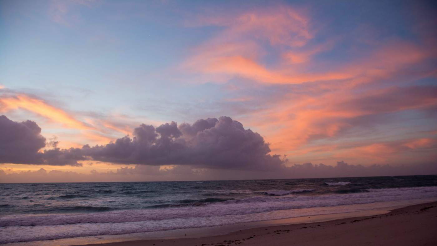 A tranquil beach at sunrise or sunset. The sky displays vibrant colors, ranging from deep purples to warm oranges and pinks. A large cumulus cloud dominates the center of the sky, casting a shadow on the horizon. Gentle waves lap onto the sandy shore. The scene evokes calmness and natural beauty.