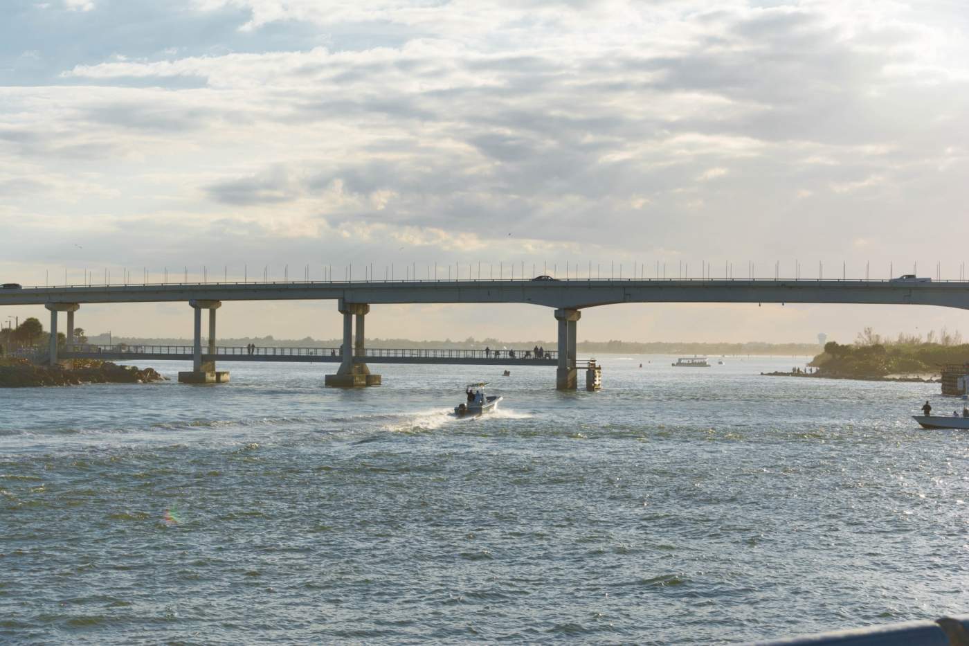 A boat navigates a wide waterway under a long concrete bridge on a partly cloudy day, with smaller boats and figures visible in the distance.