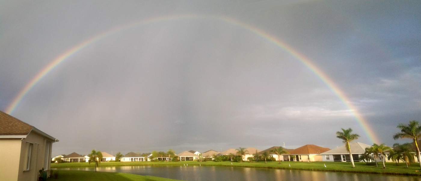 A rainbow arches over a calm residential area with houses, palm trees, and a pond under a cloudy sky.