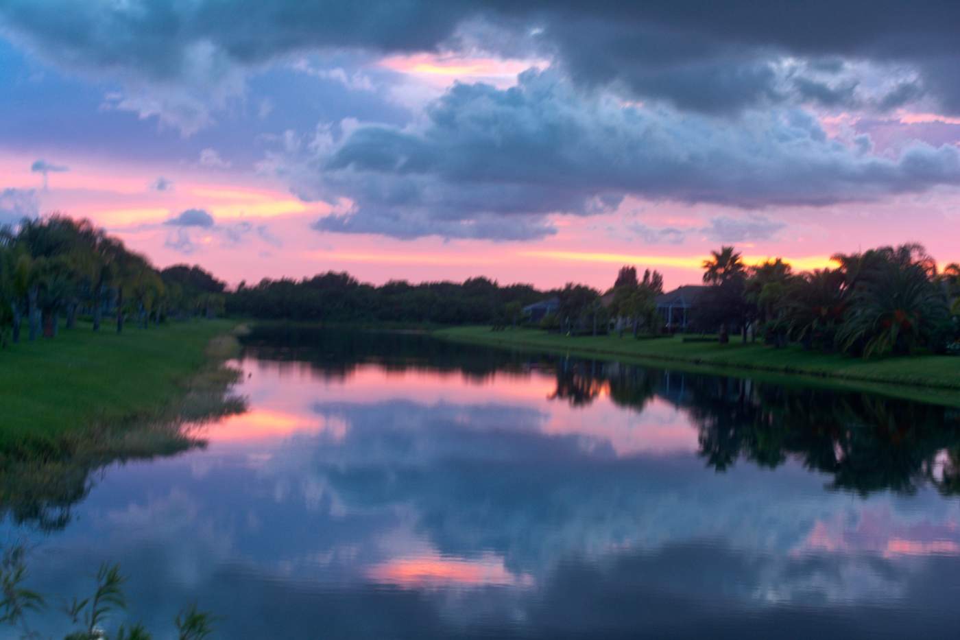 A serene lake reflects a vibrant sunset with hues of pink and purple, bordered by trees and buildings beneath a partly cloudy sky.