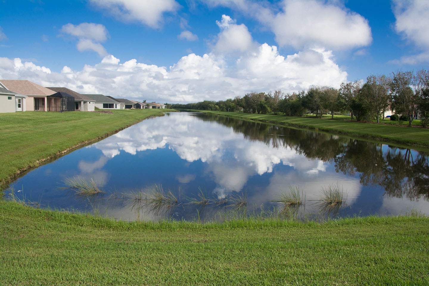 A serene pond with clear blue water reflecting the sky and clouds. Houses line one side and trees dot the other, under a partly cloudy sky.